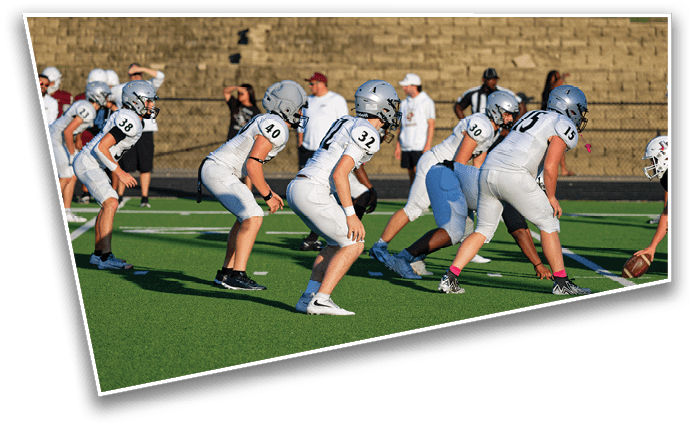 A group of people in football uniforms are lined up on a green field preparing for a play during a game. Description generated by AI