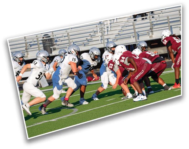 Two opposing American football teams in helmets and uniforms are lined up facing each other on a green field, ready for a play. Description generated by AI