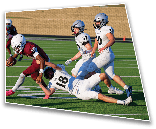 A group of people in football uniforms are actively playing on a green field with one person in a maroon uniform holding the ball while others in white uniforms attempt to tackle. Description generated by AI