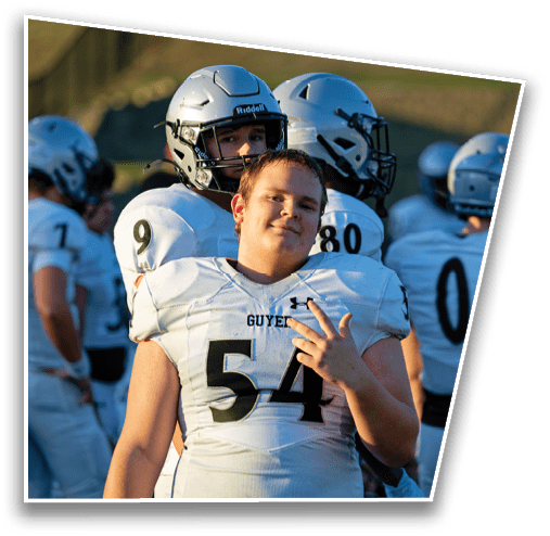 A group of people wearing white football uniforms and helmets stand together outdoors with one person in front making a hand gesture and smiling. Description generated by AI