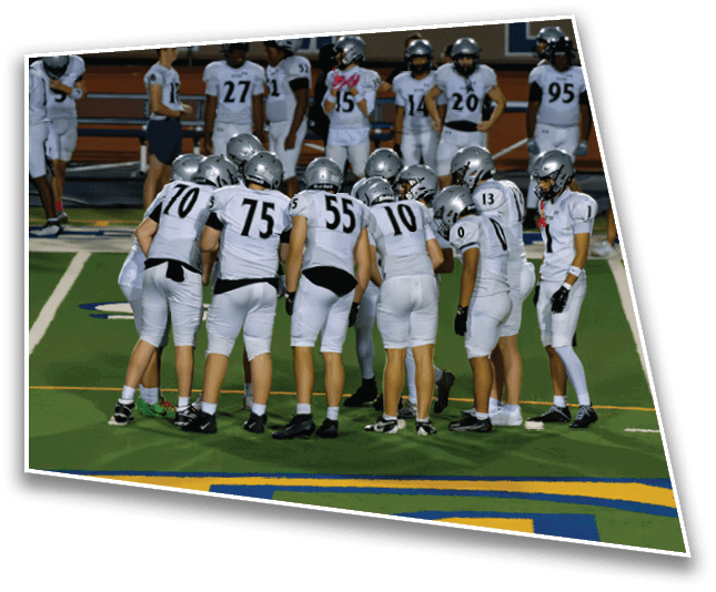 A group of people wearing white football uniforms and helmets are huddled together on a green field while others stand in the background near a fence. Description generated by AI
