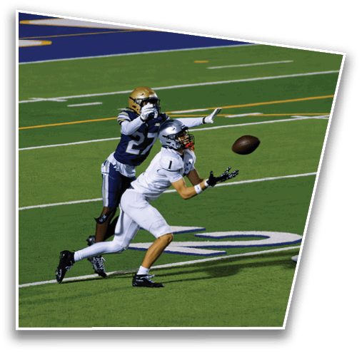 Two people in football uniforms are on a field with one person reaching out to catch a football while the other person attempts to defend. Description generated by AI