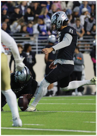 A person in a dark uniform is kicking a football held by another person kneeling on the field while a person in a white uniform stands nearby and a crowd watches from the bleachers. Description generated by AI