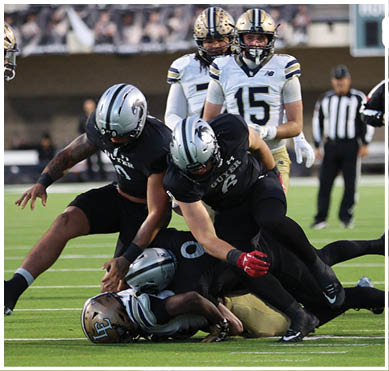 People in football uniforms are engaged in a tackle on a green field during a game. Description generated by AI