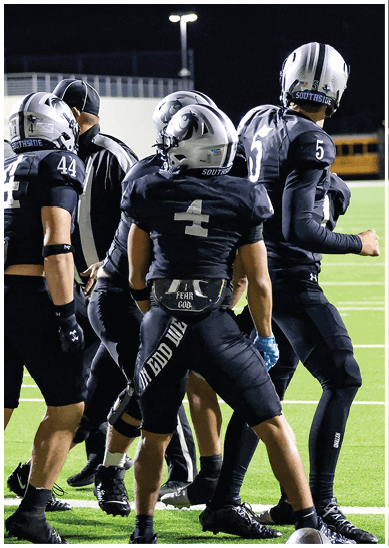 Two people in black football uniforms and helmets stand on a green field at night, with one person holding a football. Description generated by AI
