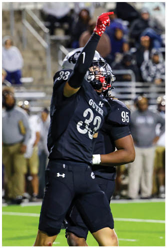 A person wearing a black football uniform with the number 33 is raising one arm while standing on a football field with another player in the background. Description generated by AI