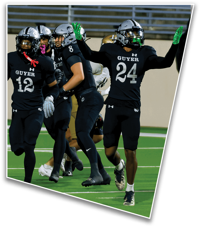 Two people wearing football helmets and black jerseys with the word Guyer are on a field, with one raising a green-gloved hand. Description generated by AI