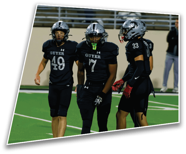 Four people in black football uniforms and helmets stand on a green field with one person in casual clothing in the background. Description generated by AI