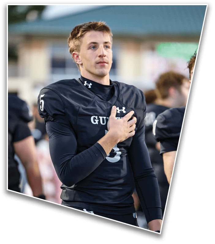 A person in a black football uniform with the number 5 on the sleeve stands with one hand over their chest, appearing focused during a pre-game or national anthem moment. Description generated by AI