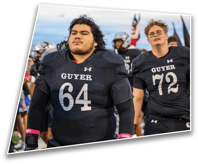 Two people wearing black football uniforms with the word “GUYER" and numbers on the front stand on a field with other people and players in the background. Description generated by AI