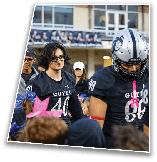 People wearing football uniforms and casual clothing are gathered outdoors near a building, with some children wearing colorful bows in their hair. Description generated by AI