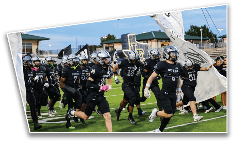 A group of people in black sports uniforms and helmets are running onto a football field through a large banner. Description generated by AI