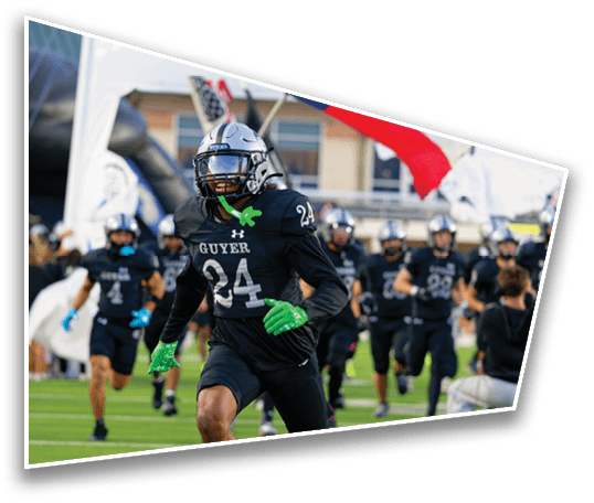 A person wearing a black football uniform with the number 24 and green gloves is running on a field with a group of similarly dressed players and flags in the background. Description generated by AI