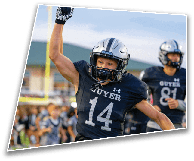 A person wearing a football uniform with the number 14 and a helmet is raising one arm in celebration on a football field with other players and spectators in the background. Description generated by AI