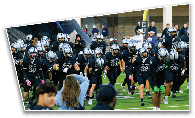 A group of football players in black uniforms and silver helmets are running onto a field with spectators in the foreground. Description generated by AI