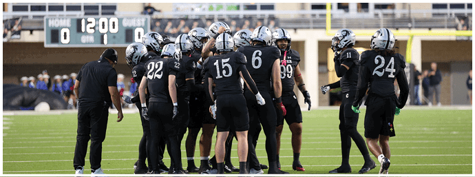 A group of people wearing black sports uniforms and helmets are huddled together on a football field near a scoreboard showing a tied score. Description generated by AI