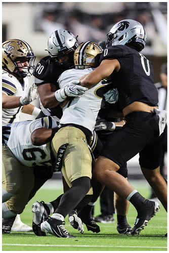 Several people in football uniforms are engaged in a close physical tackle on a grassy field during a game. Description generated by AI