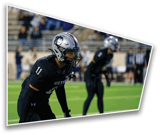 Two people in black football uniforms and silver helmets are positioned on a green field with blurred spectators in the background. Description generated by AI