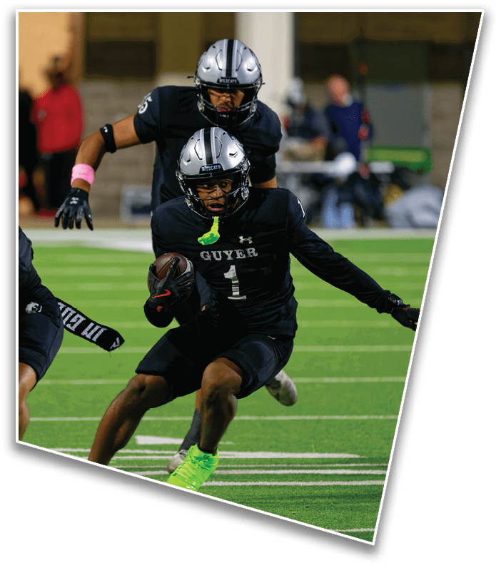 Two people in black football uniforms with helmets are playing on a green field, with one holding the ball and running while the other follows closely behind. Description generated by AI