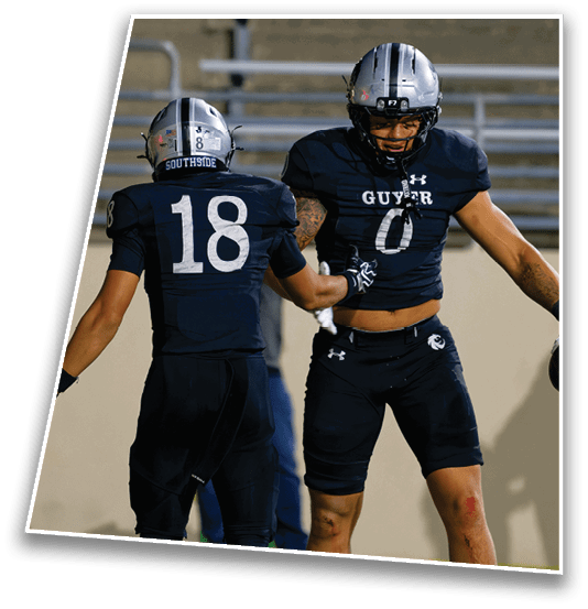 Two football players in dark uniforms and helmets celebrate on the field with one holding a football. Description generated by AI