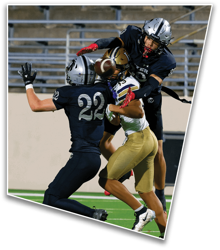 Three football players in action on a field, with two players in black uniforms attempting to block or tackle a player in a white and gold uniform who is holding the ball. Description generated by AI