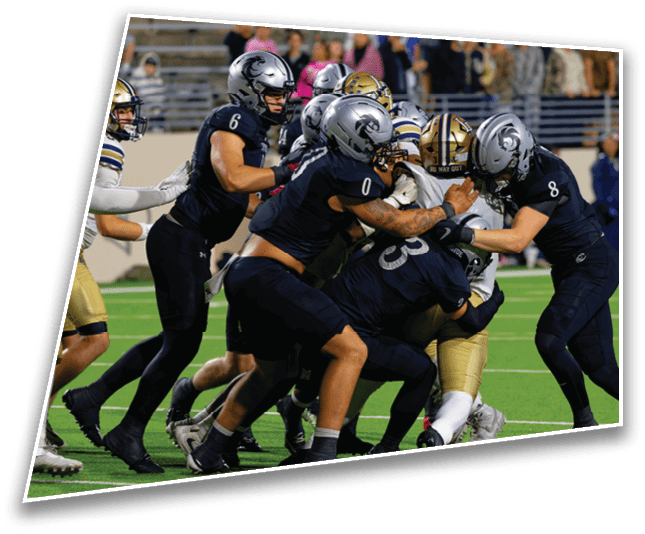 Several football players in dark and gold uniforms are engaged in a close tackle on a green field during a game. Description generated by AI
