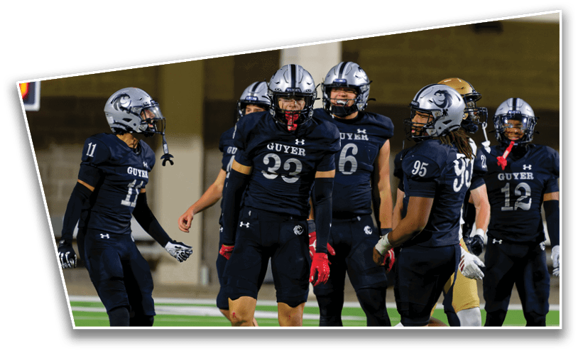 A group of football players in dark uniforms and helmets are gathered on a field, appearing to communicate or strategize during a game. Description generated by AI