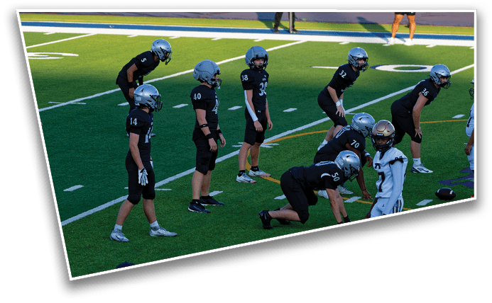 A group of football players in black uniforms and helmets are lined up on a green field preparing for a play against an opposing player in a white uniform. Description generated by AI