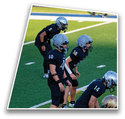 Four people in football uniforms and helmets are positioned on a green field, appearing ready for a play. Description generated by AI