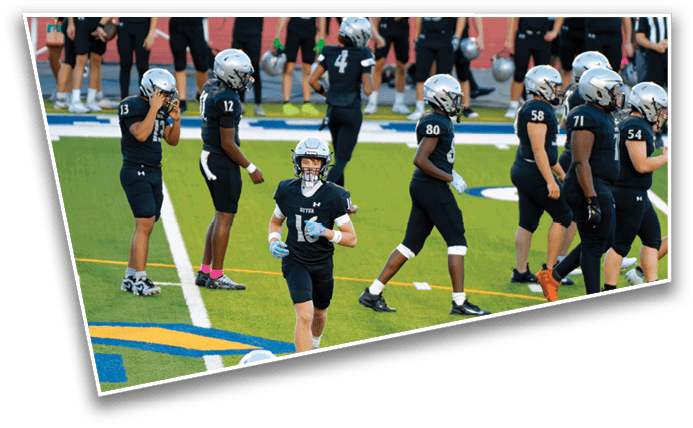 People wearing football uniforms and helmets are standing and walking on a green field with white yard lines and a blue and yellow logo. Description generated by AI