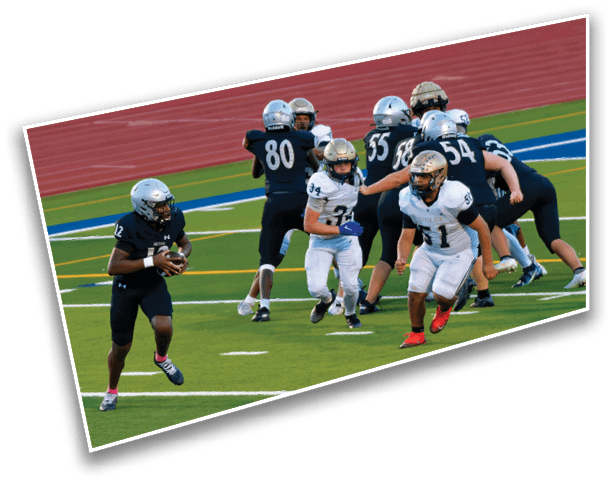People wearing football uniforms are engaged in a play on a green field with a red running track in the background. Description generated by AI