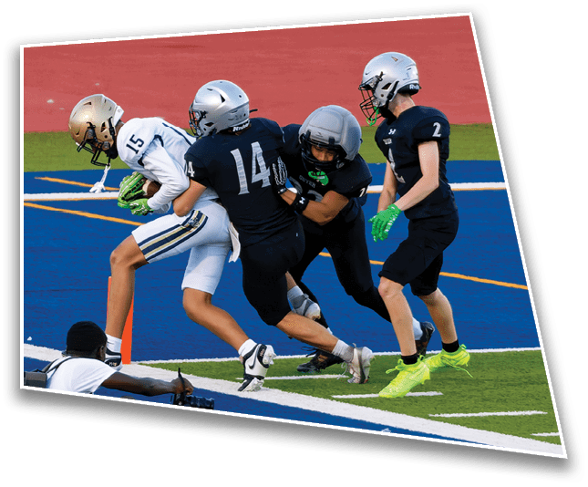 Four people in football uniforms are engaged in a play on a field with one person holding the ball while another person with a camera is sitting on the ground nearby. Description generated by AI