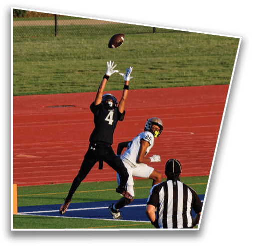 A person wearing a helmet and gloves is reaching up to catch a football in an outdoor sports field with a red track and green grass. Description generated by AI