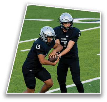 Two people wearing football helmets and dark jerseys with the word “GUYER" are on a green field, appearing focused during a game or practice. Description generated by AI
