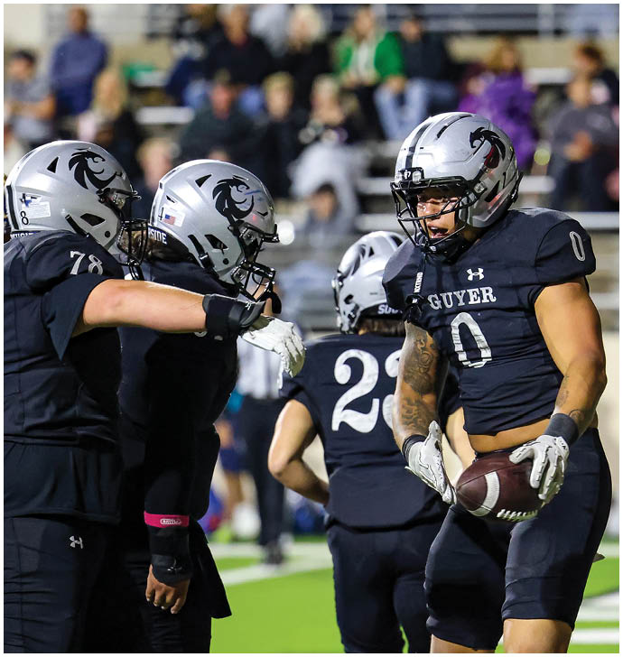 Four football players in black uniforms and silver helmets are on a field with one holding a football while the others appear to be communicating or celebrating. Description generated by AI