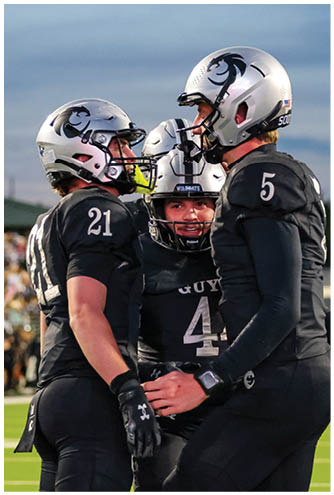 Three people wearing black football uniforms and silver helmets are standing close together on a field with a crowd in the background. Description generated by AI
