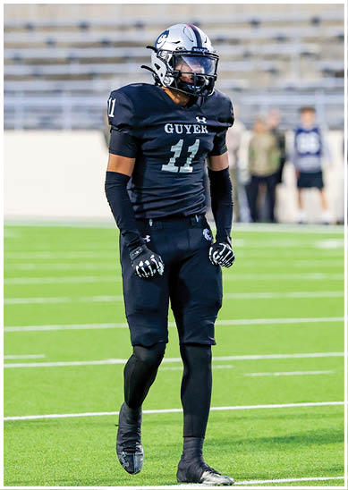 A person wearing a black football uniform with the number 11 and the word “GUYER" stands on a green football field with empty bleachers in the background. Description generated by AI