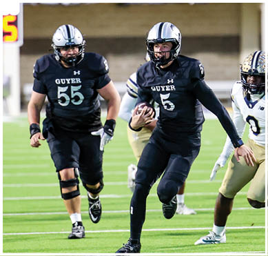A person in a black football uniform with the number 5 is running with the ball while being chased by a person in a white and gold football uniform with the number 0 on a green field. Description generated by AI
