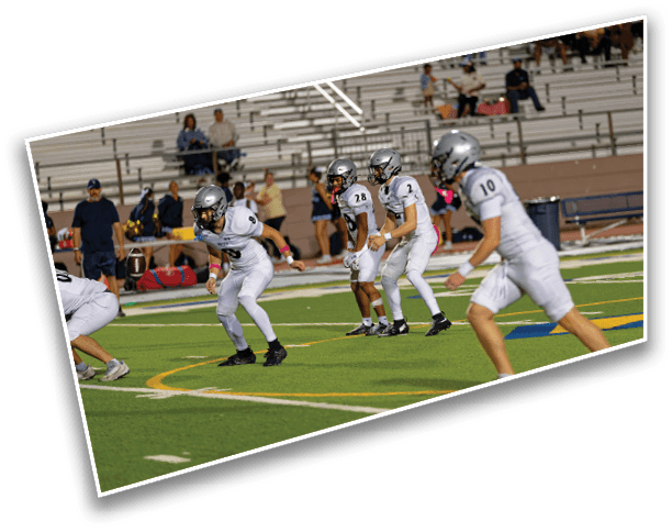 Four people in white football uniforms and helmets are positioned on a football field preparing for a play with empty bleachers and a few spectators in the background. Description generated by AI
