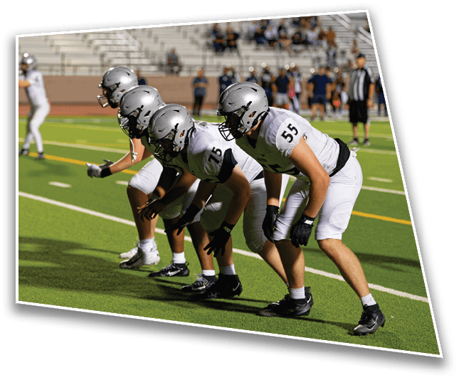 A group of people in football uniforms and helmets are lined up on a field in a ready stance while one person in the background prepares to throw a football. Description generated by AI