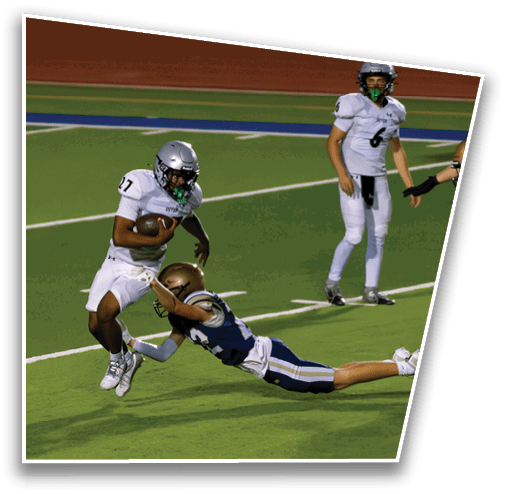 A person in white football gear is running with the ball while another person in blue football gear dives attempting to tackle them on a green field. Description generated by AI