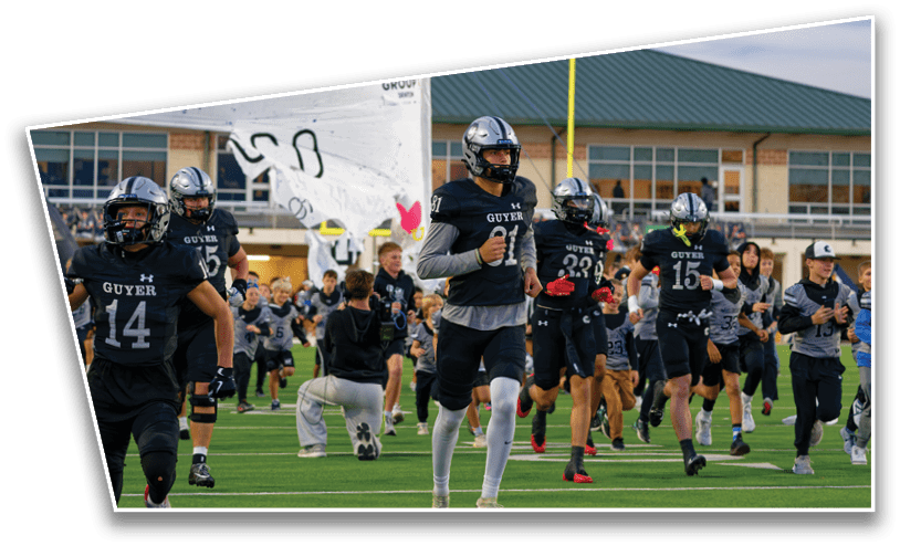 A group of people in football uniforms are running onto a field through a banner with a large dog graphic in front of a stadium. Description generated by AI
