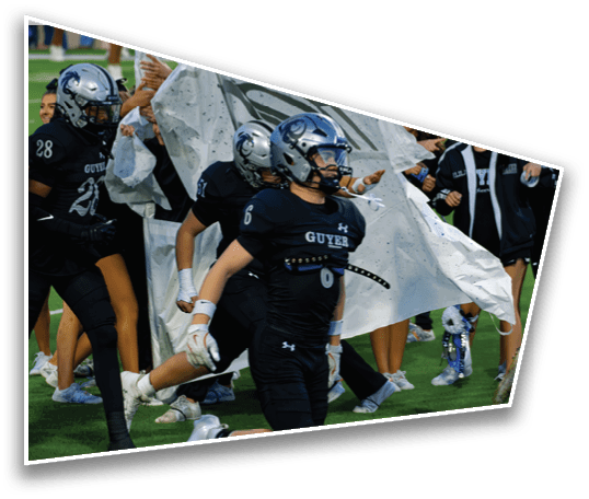 People in football uniforms are running onto a field through a torn banner held by others in casual clothing. Description generated by AI