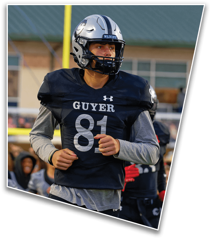 A person wearing a silver football helmet with a black faceguard and a black jersey is looking to the side on a football field with a goalpost and a green-roofed building in the background. Description generated by AI