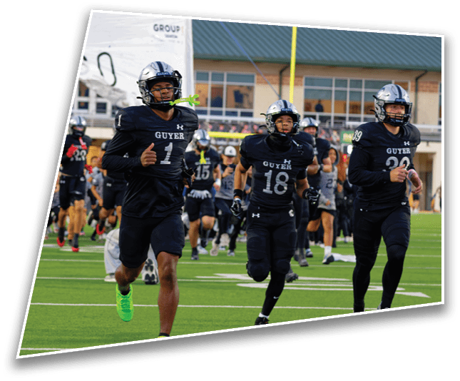 People wearing black football uniforms with helmets are running onto a green field from behind a torn white banner in front of a building with large windows. Description generated by AI