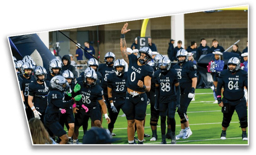 A group of football players in black uniforms and helmets are gathered on a field with one player raising an arm while others stand around. Description generated by AI