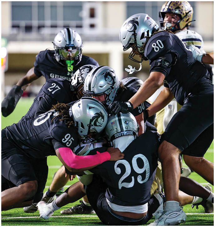 Several football players in black uniforms and helmets are tackling a player in a white uniform on a grassy field during a game. Description generated by AI