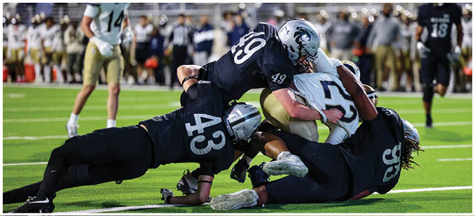Three football players in dark uniforms tackle a player in a white and gold uniform on a green field during a game. Description generated by AI