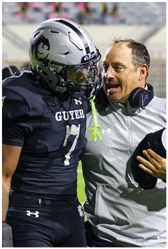 A person wearing a headset and light-colored jacket is talking closely to a football player in a black uniform and helmet on a sports field. Description generated by AI