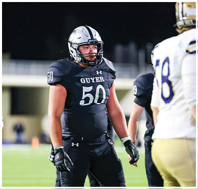 A person wearing a black football uniform with the number 50 and the word Guyer stands on a football field at night while other players and a referee are nearby. Description generated by AI